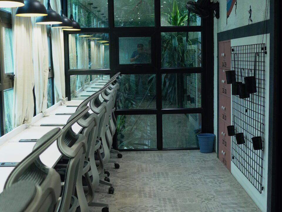 Row of modern ergonomic chairs and white desks in a well-lit coworking space with large glass windows, hanging black lamps, and a green outdoor view.